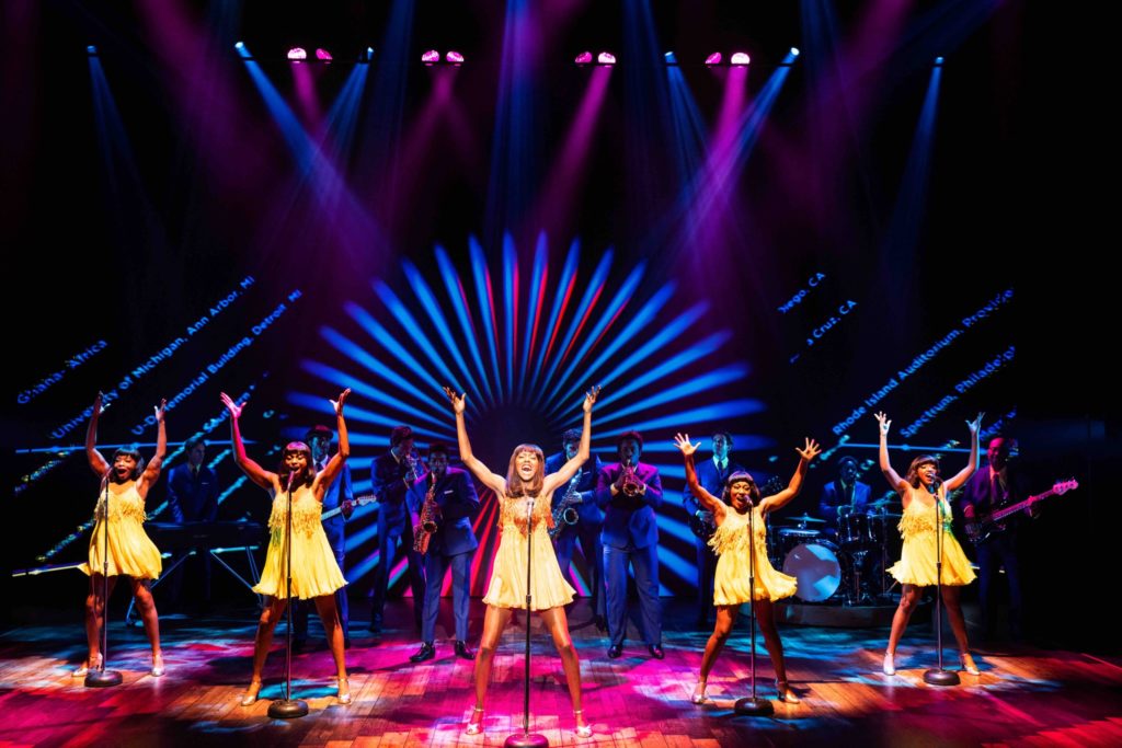 The cast of Tina: The Tina Turner Musical sings and dances on a light-filled stage in bright yellow dresses during the song "I Want To Take You Higher."